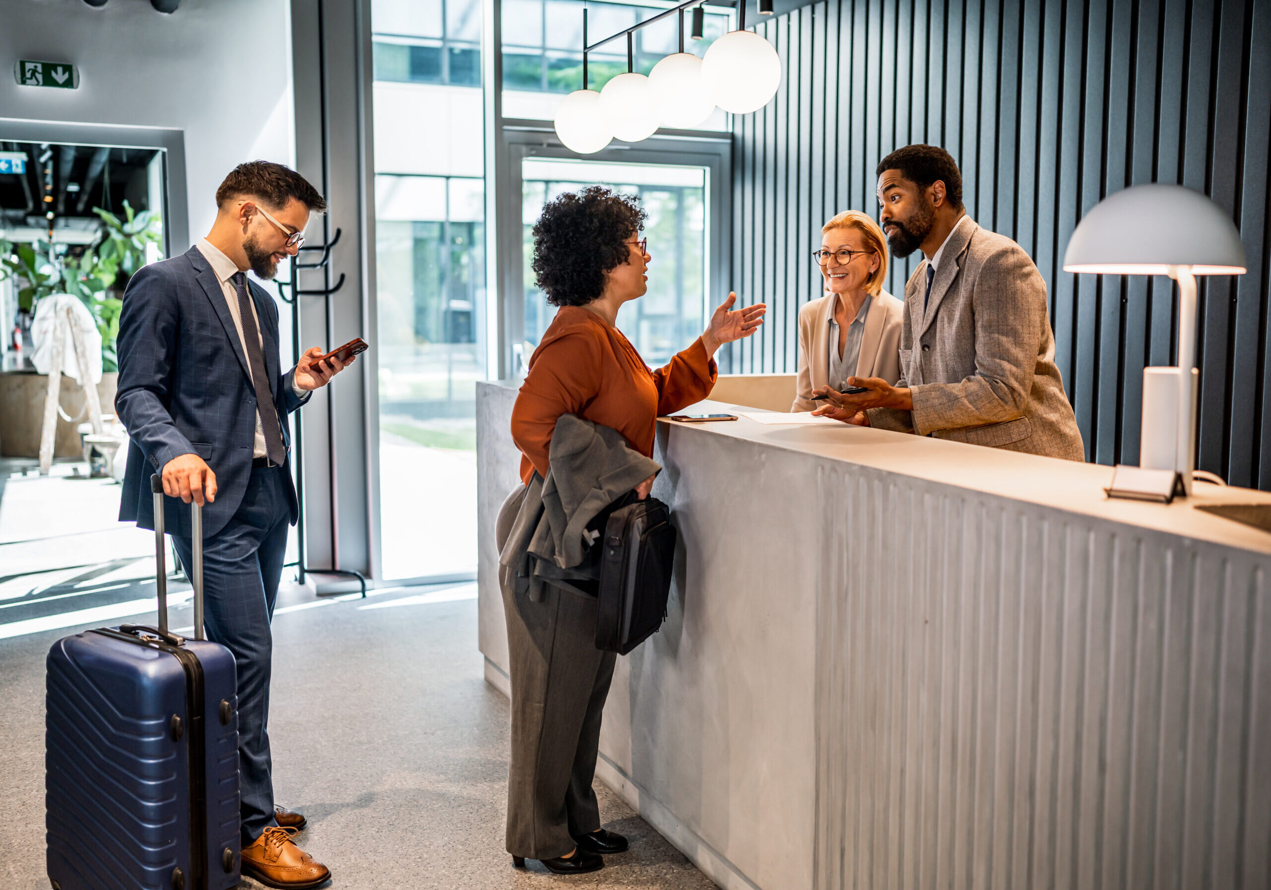 Businesswoman talking to receptionist during check in process at hotel reception desk, businessman waiting with suitcase